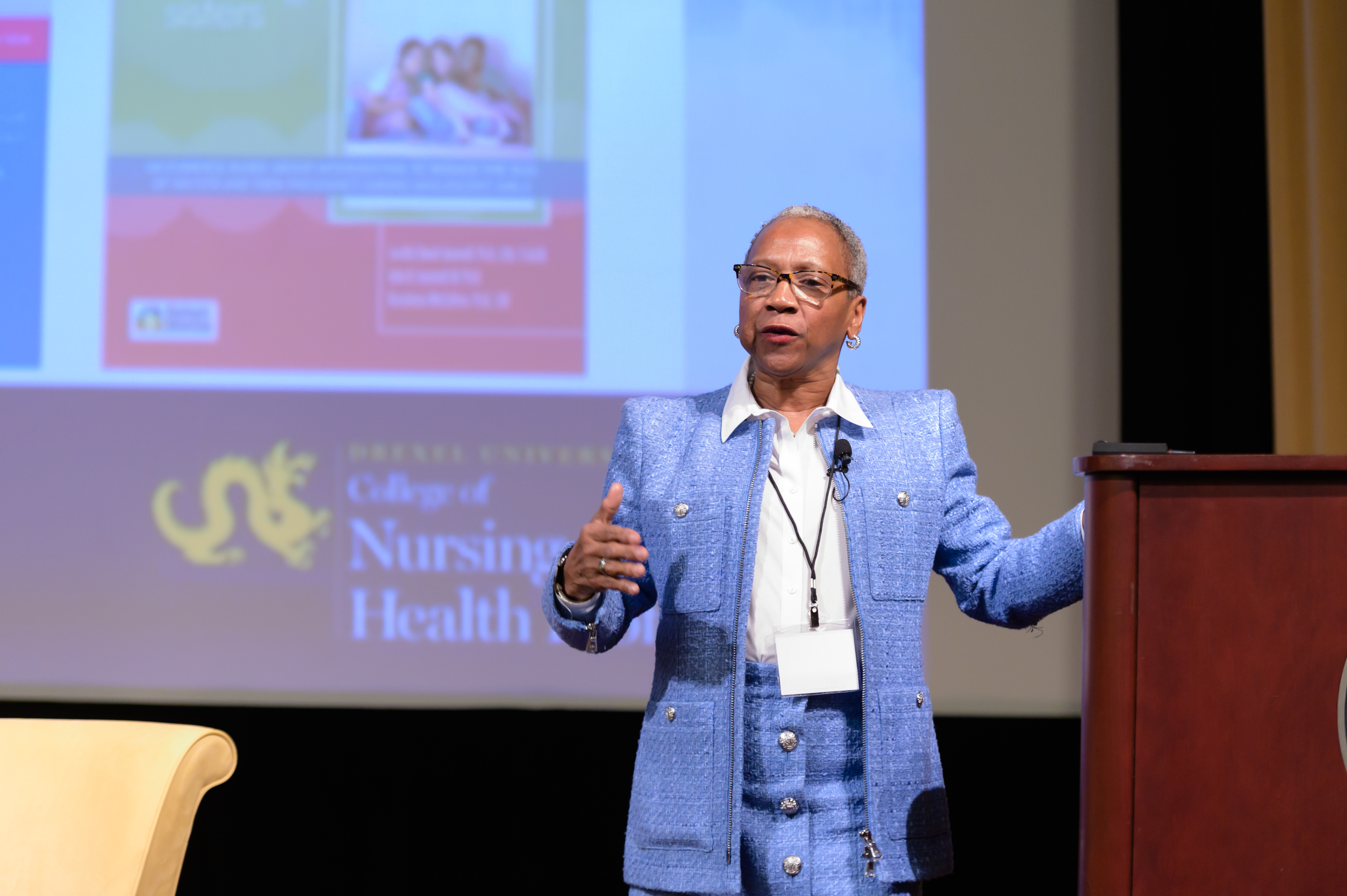 A woman wearing a blue suit is speaking at a podium in a formal setting, possibly a conference or seminar. Behind her is a projection screen displaying a presentation with the logo of Drexel University College of Nursing and Health Professions.