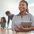 Professional looking young woman standing in a conference room while smiling and holding a tablet, with a group of people working in the background.