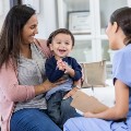 A smiling parent sits in an exam room holding her happy child and talking with a health care provider